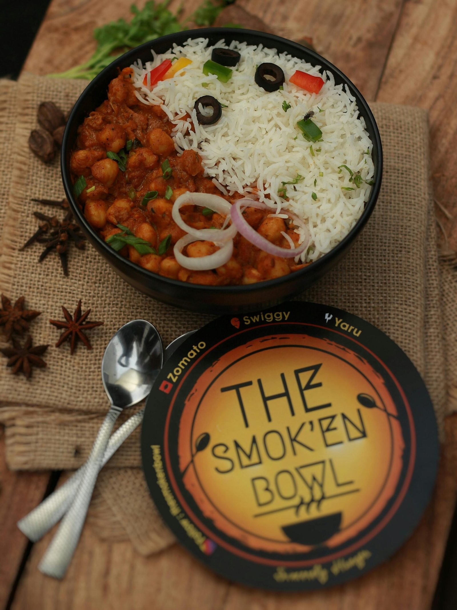 Spicy Indian curry with rice and vegetables in a black bowl, served with a side of spices.